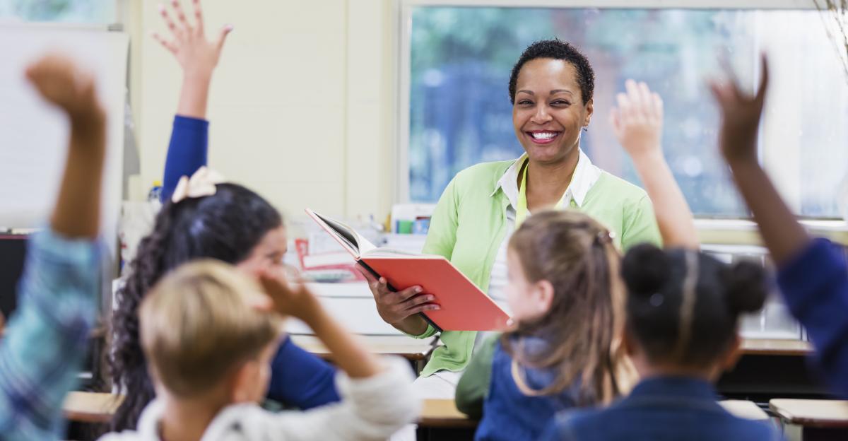 An African-American teacher, a mature woman in her 40s, sitting in front of her class of elementary school students, 6 and 7 years old, reading a book. She has asked a question and most of the children are raising their hands. They are in first grade or second grade.