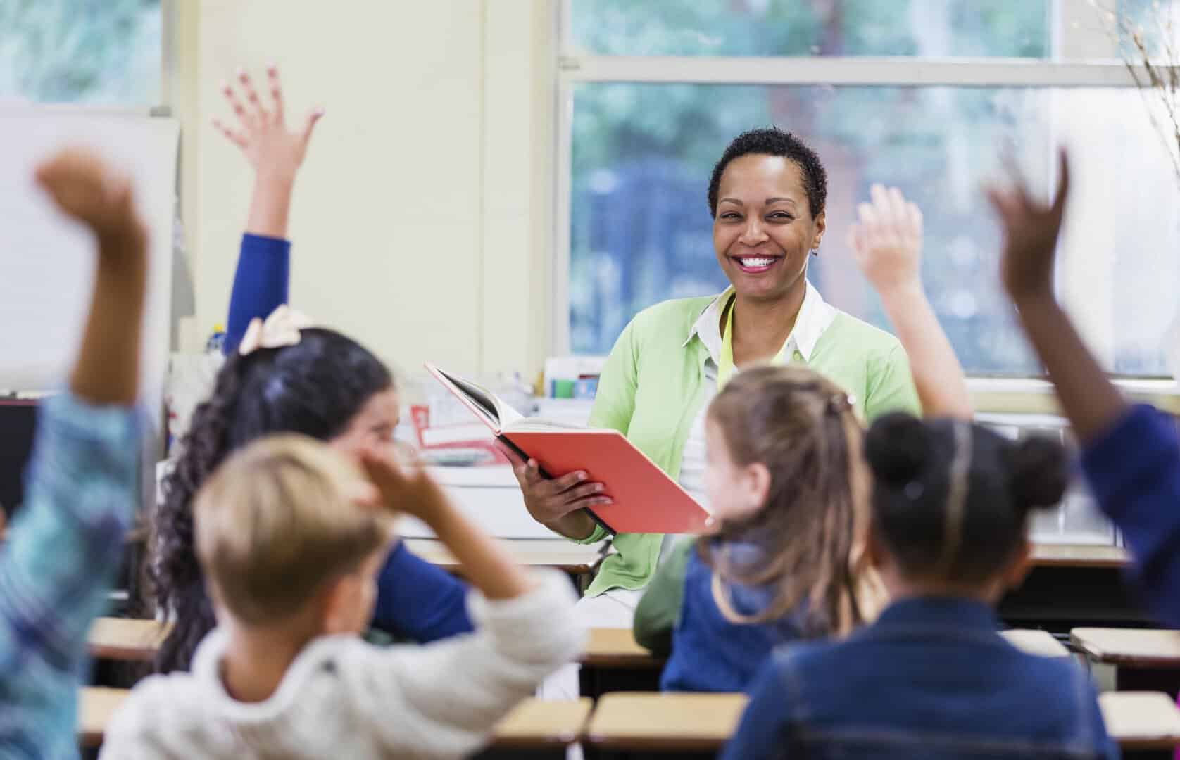 An African-American teacher, a mature woman in her 40s, sitting in front of her class of elementary school students, 6 and 7 years old, reading a book. She has asked a question and most of the children are raising their hands. They are in first grade or second grade.