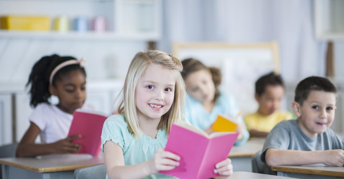A preschool student sits at a desk in her classroom with an open book. She is smiling at the camera.