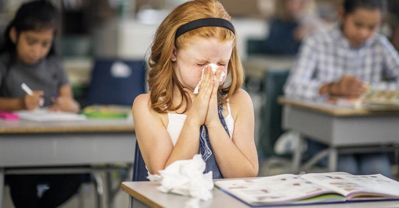 young student with cold in classroom blowing her nose into facial tissue