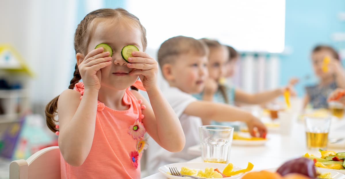 kids eating nutritious food in the classroom centered on young funny girl holding cucumber slices over her eyes