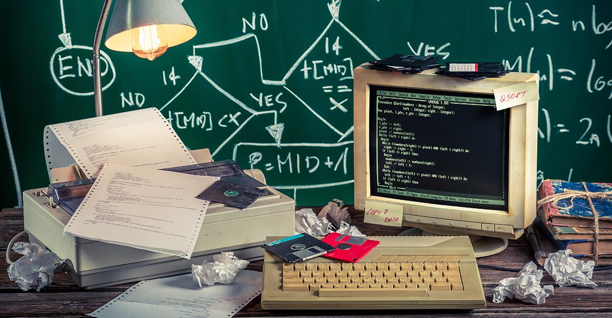 teacher's desk with an old pc, printer, and other pieces of outdated tech; background is a green chalkboard with formulas written on it