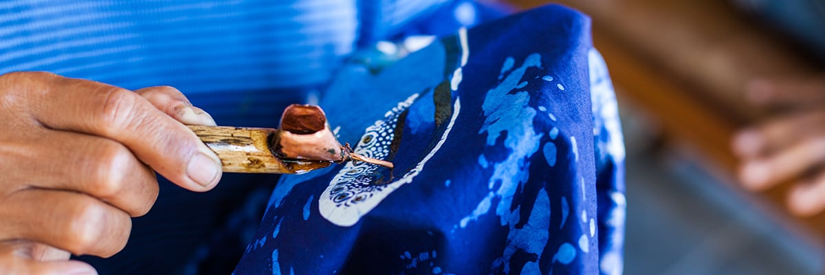 artist using a homemade wax applicator to create a batik art design on blue fabric