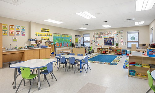 early childhood classroom with colorful rugs and decorations, wobble stools, and tables and chairs, full room view