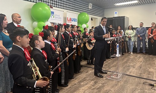man in a suit speaking at a podium with other educators and a school mariachi band standing behind him