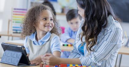 young student using a digital tablet and math manipulatives to practice math, smiling up at teacher