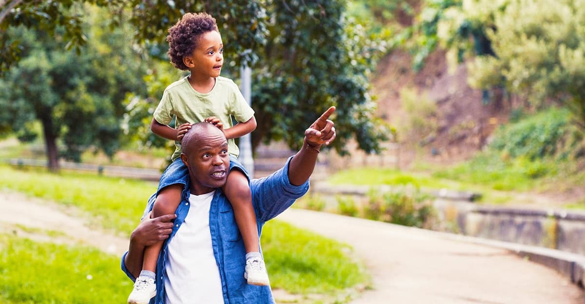 father pointing to something in the distance, his son is riding piggyback and looking in the direction he's pointing