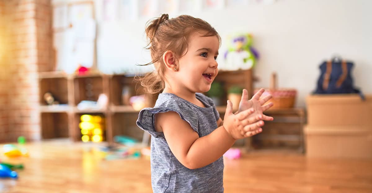 early childhood toddler clapping hands in classroom, blurry sensory tools scattered in background