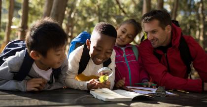 teacher and students outside, one student looking at a leaf through a magnifying glass and taking notes