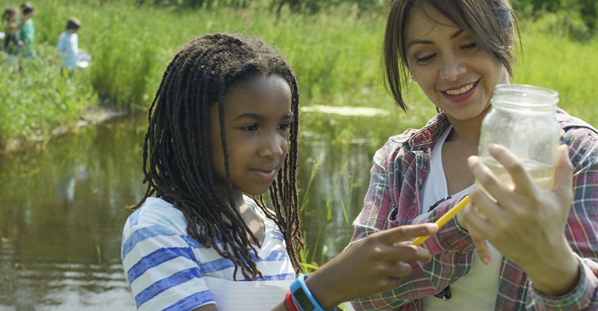 student and teacher looking at water in a jar, pond and tall grass in the background