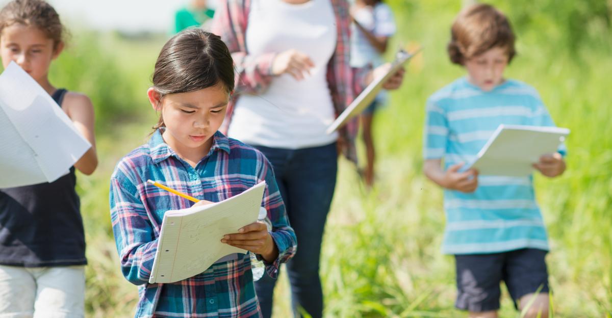 An elementary school girl of Filipino ethnicity is outdoors with her class exploring nature. She is busy writing on her notepad while walking with the group.