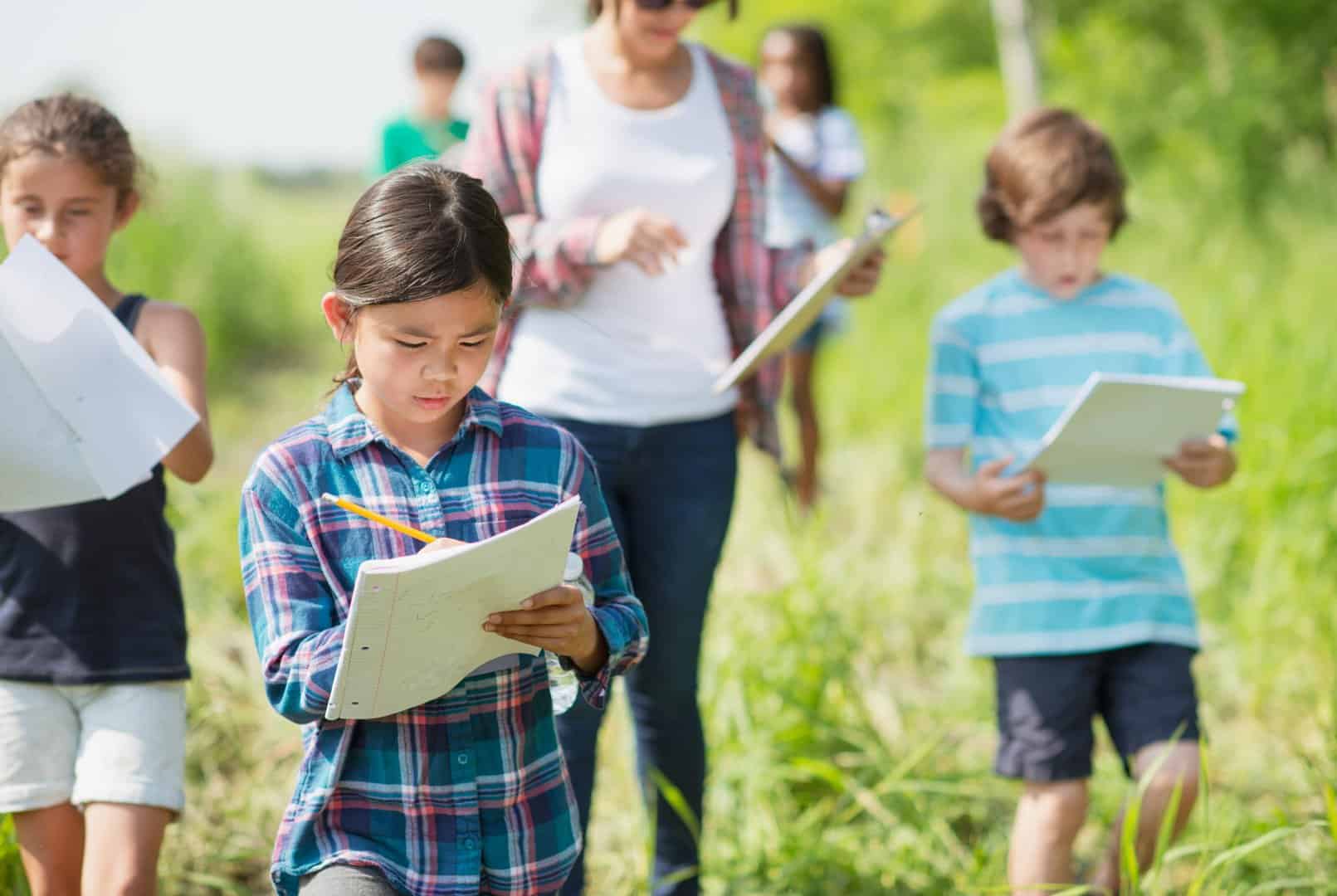 An elementary school girl of Filipino ethnicity is outdoors with her class exploring nature. She is busy writing on her notepad while walking with the group.