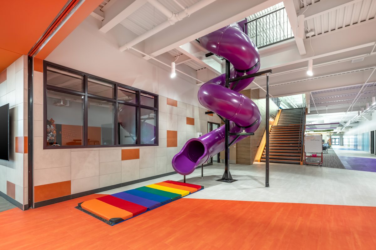 An indoor purple corkscrew enclosed slide that ends above a rainbow mat in a hallway in a school.