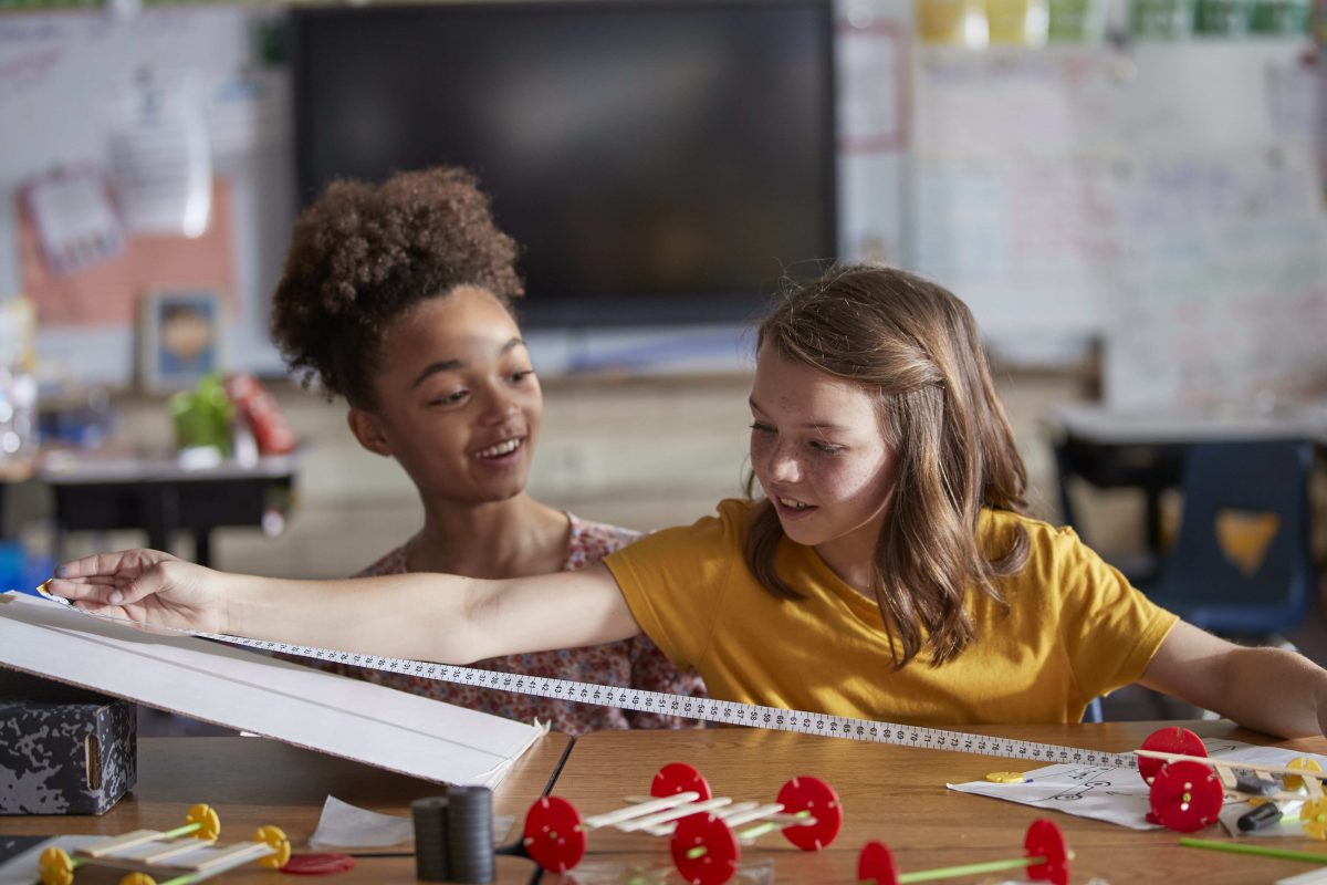 Two female students in a classroom. One is holding a measuring tape.