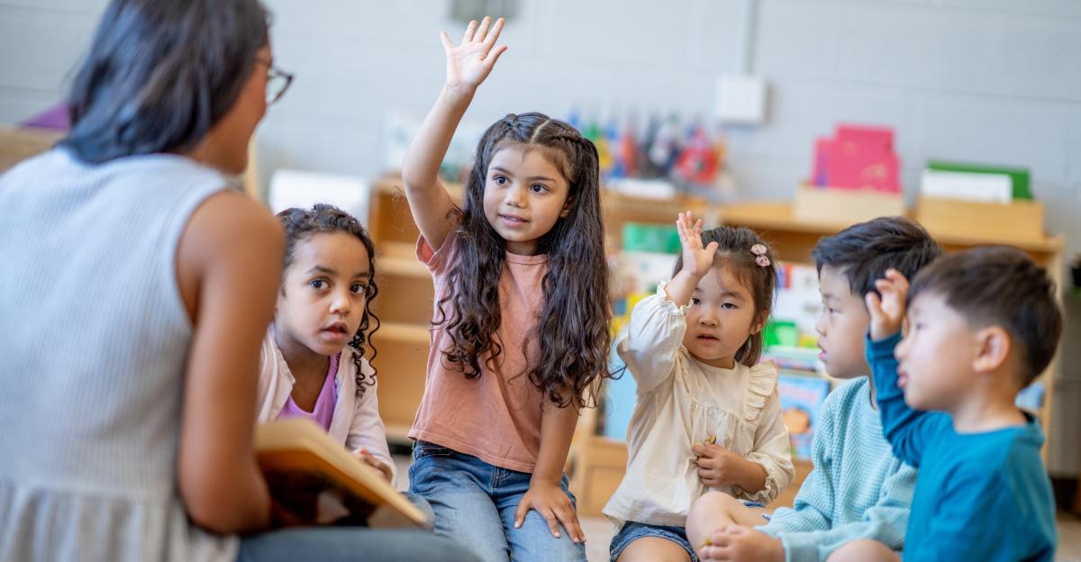 A female Montessori teacher sits in front of a small group of children as she reads them a story. The children are each dressed casually and are listening attentively.
