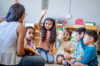 A female Montessori teacher sits in front of a small group of children as she reads them a story. The children are each dressed casually and are listening attentively.