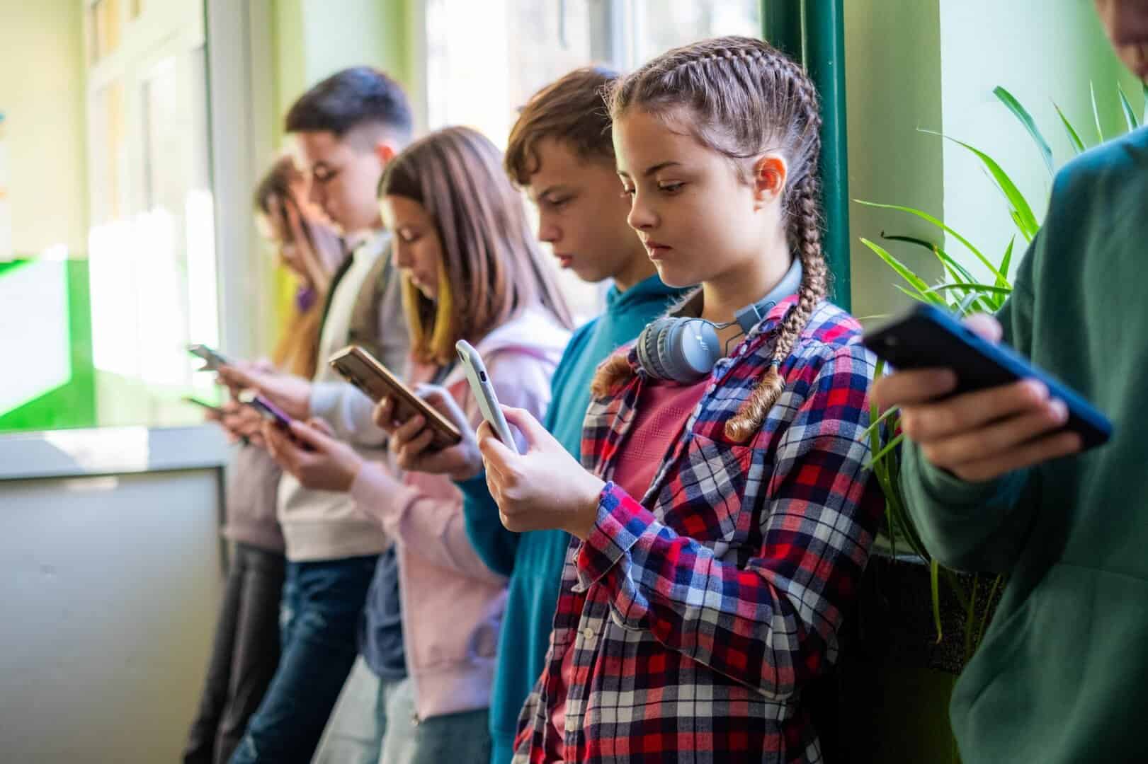 Teenage students are standing in the school hallway, all looking at their phones.