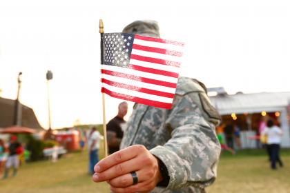 U.S. veteran holding American flag. Anonymous army solider in National Guard wearing military uniform. Freedom concept for armed forces, military pride, patriotism on independence day / July 4th / Fourth of July or Veteran's Day. Waist up candid portrait or real life real people outdoors during summertime at a county fair in America.