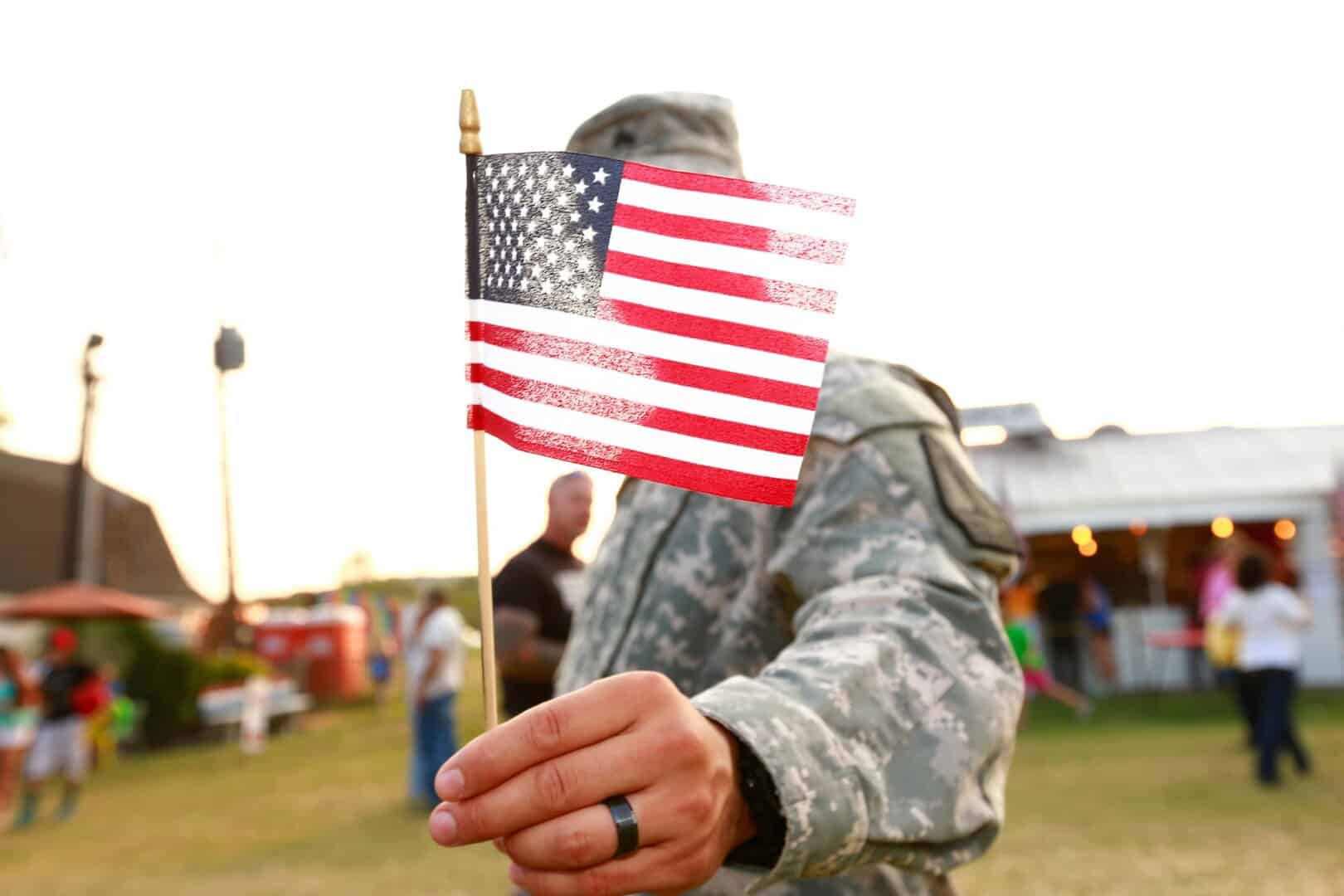 U.S. veteran holding American flag. Anonymous army solider in National Guard wearing military uniform. Freedom concept for armed forces, military pride, patriotism on independence day / July 4th / Fourth of July or Veteran's Day. Waist up candid portrait or real life real people outdoors during summertime at a county fair in America.
