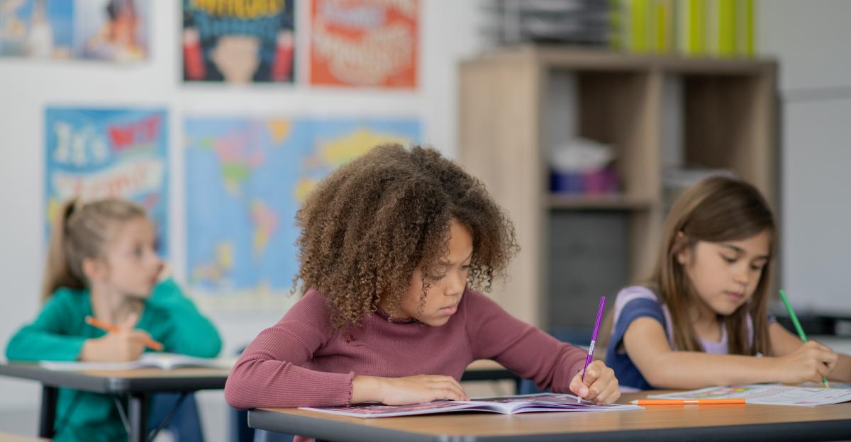 A young elementary student sits at her desk among her peers, as she works away independently. She is dressed casually and focused on her work.