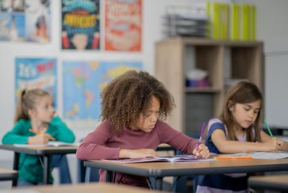 A young elementary student sits at her desk among her peers, as she works away independently. She is dressed casually and focused on her work.