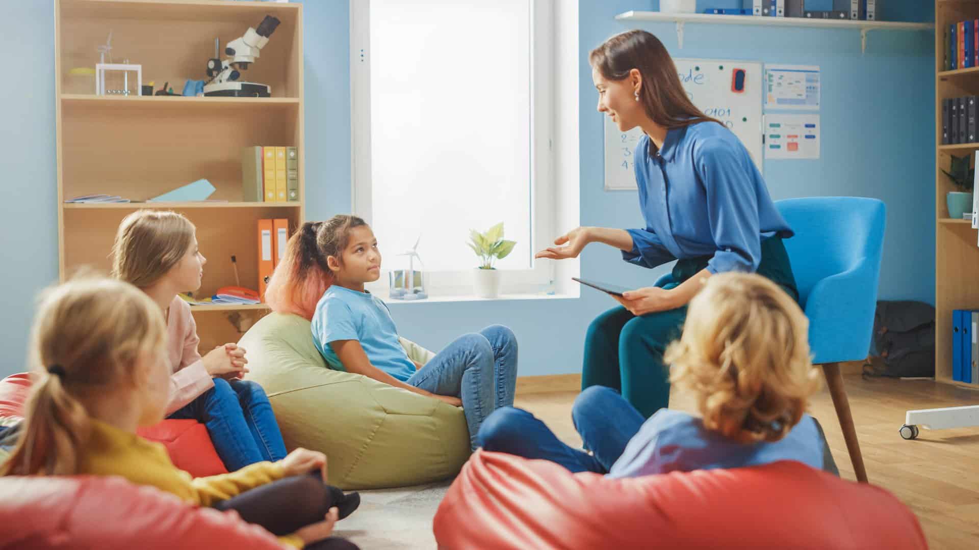 School Creativity Class: Children Sitting on the Bean Bags while Caring Teacher Explains Lesson while Using Digital Tablet Computer. Smart Children Learning in Friendly Modern Environment.