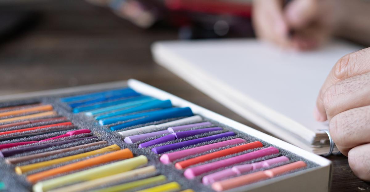 person, artist painting with a pastel crayon chalk on a white sheet of paper and pellette of pastel chalks in front of him on the wooden table, selective focus