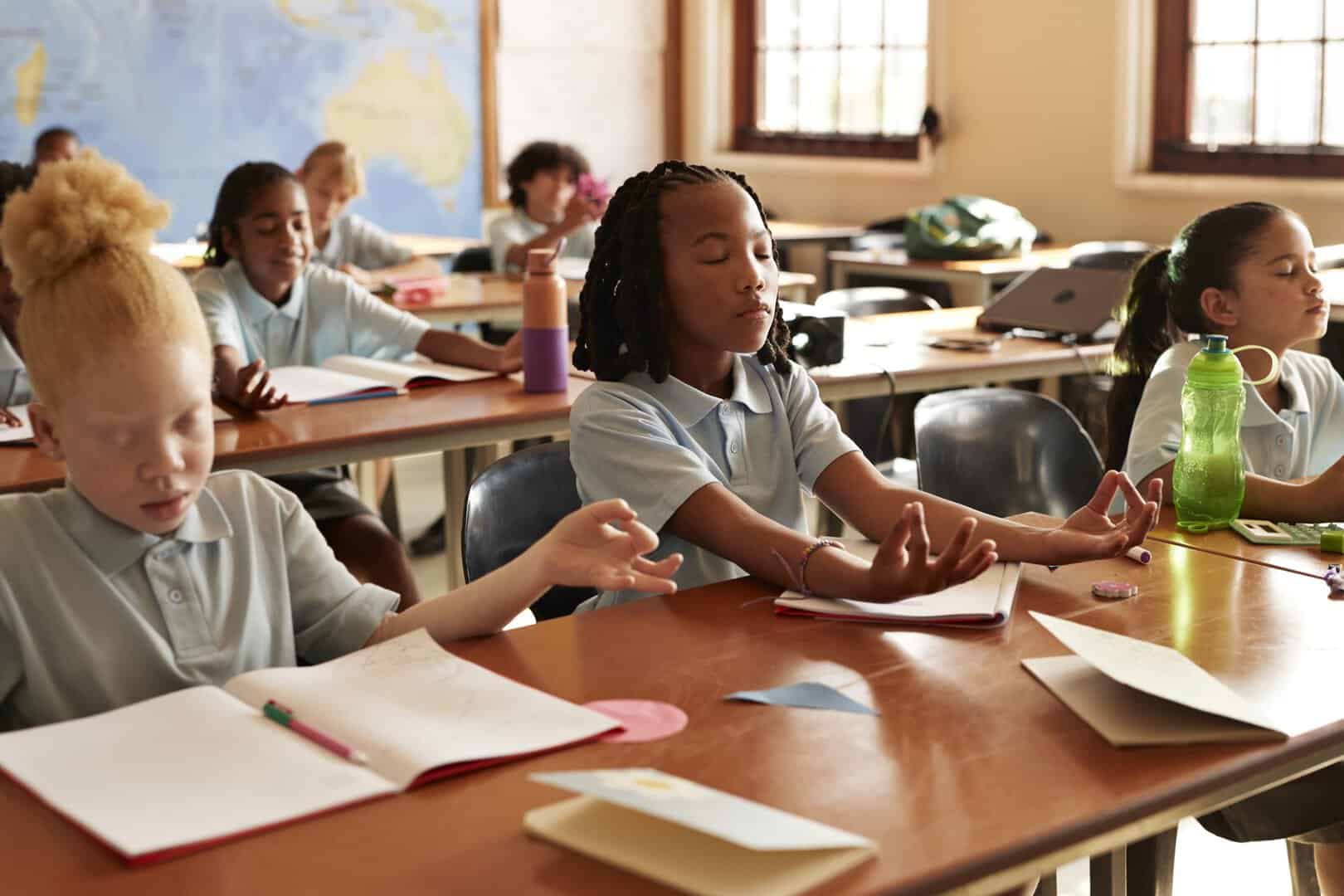 Group of multiracial students meditating while sitting at desks in classroom