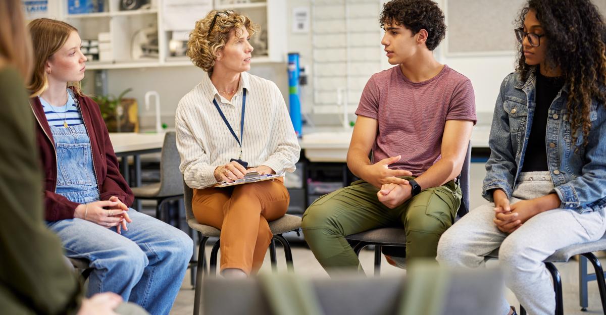 Multiethnic teenagers sitting in a circle engaged in an open group conversation to psychological support person. Instructor leading a thoughtful discussion with students in a relaxed setting. Mature woman supporting a group of girls and guys during a group therapy meeting session, giving advice.